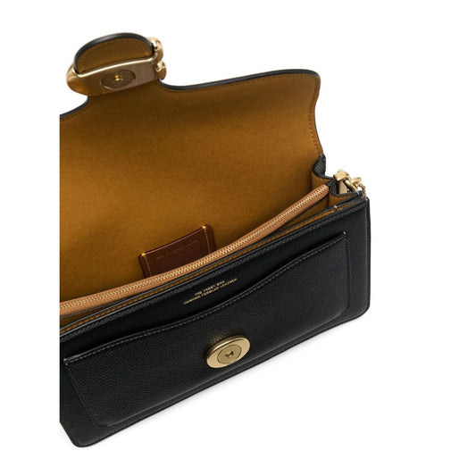 Model holding Coach Tabby 26 leather shoulder bag in earth brown with pebbled texture and logo plaque, detachable handle, and shoulder strap.